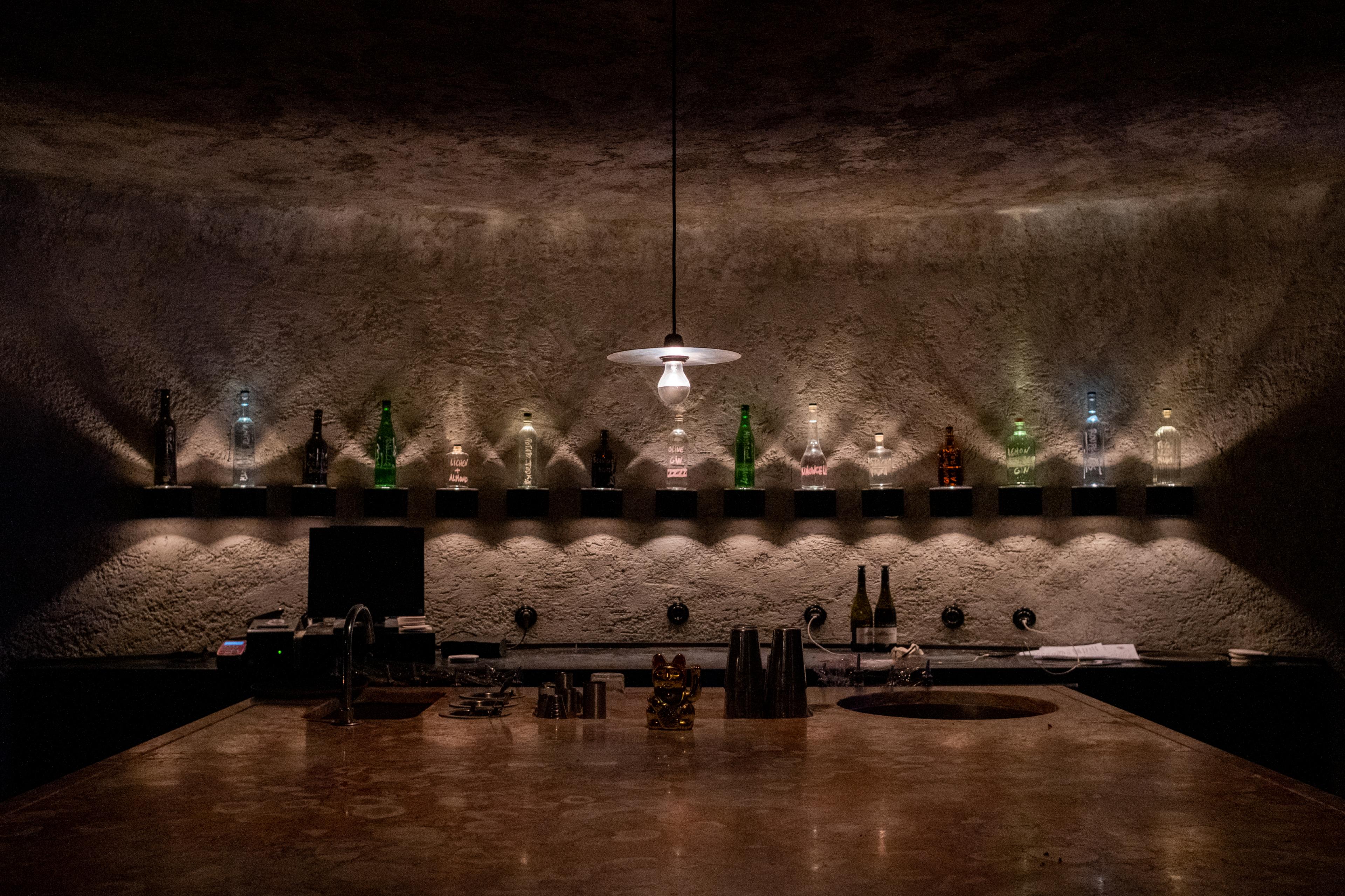 A single pendant light illuminates a pink limestone bar and up-lit shelf lined with various liquor bottles.
