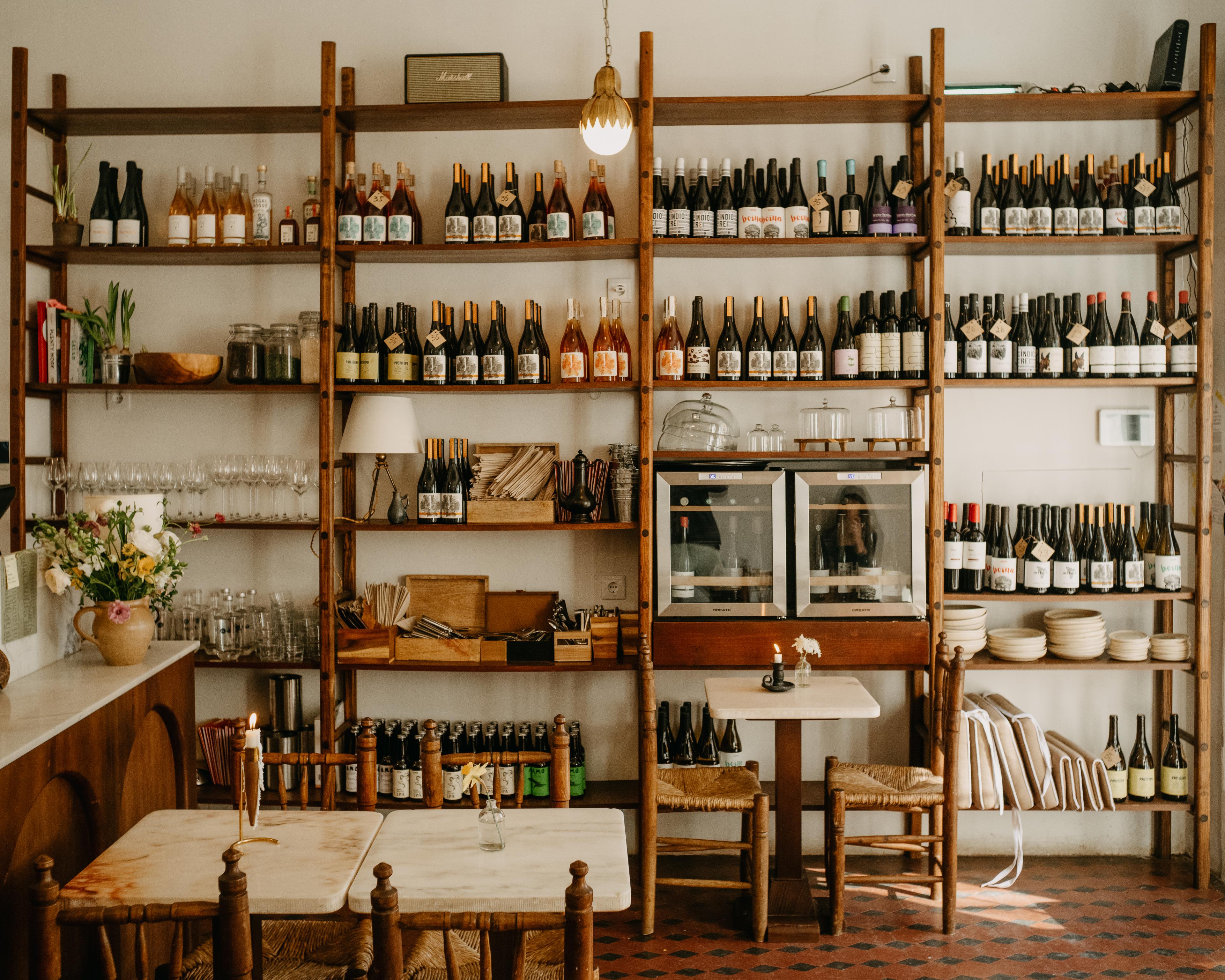 Wooden chairs and square tables sit beside a restaurant wall covered in dark shelving lined with wine bottles. 