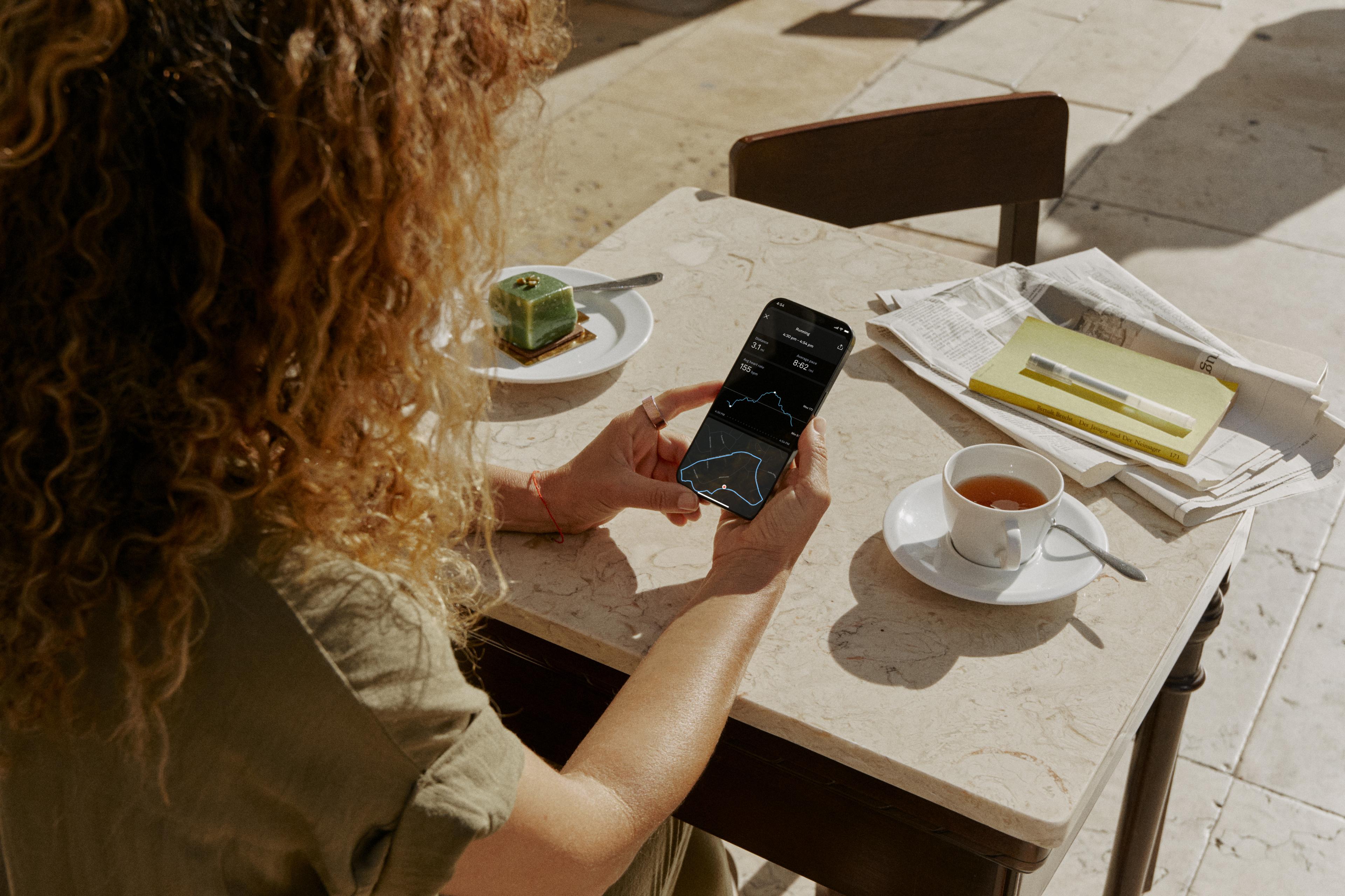 Person checking health data on a smartphone while wearing an oura ring at a café table with tea and pastry