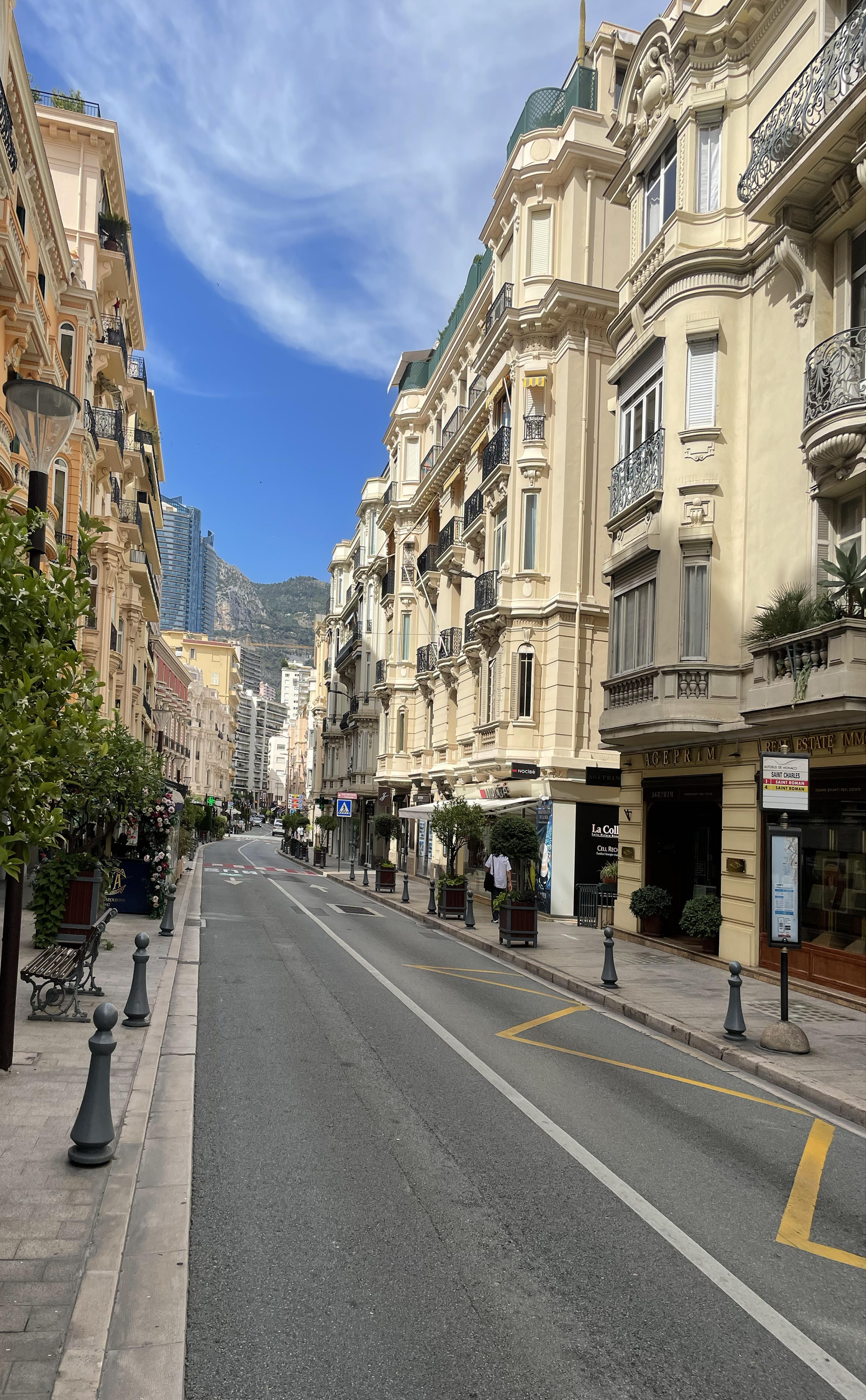 Elegant Belle Époque street with cream-colored ornamental facades, narrow yellow-marked road, and mountains visible in the distance.