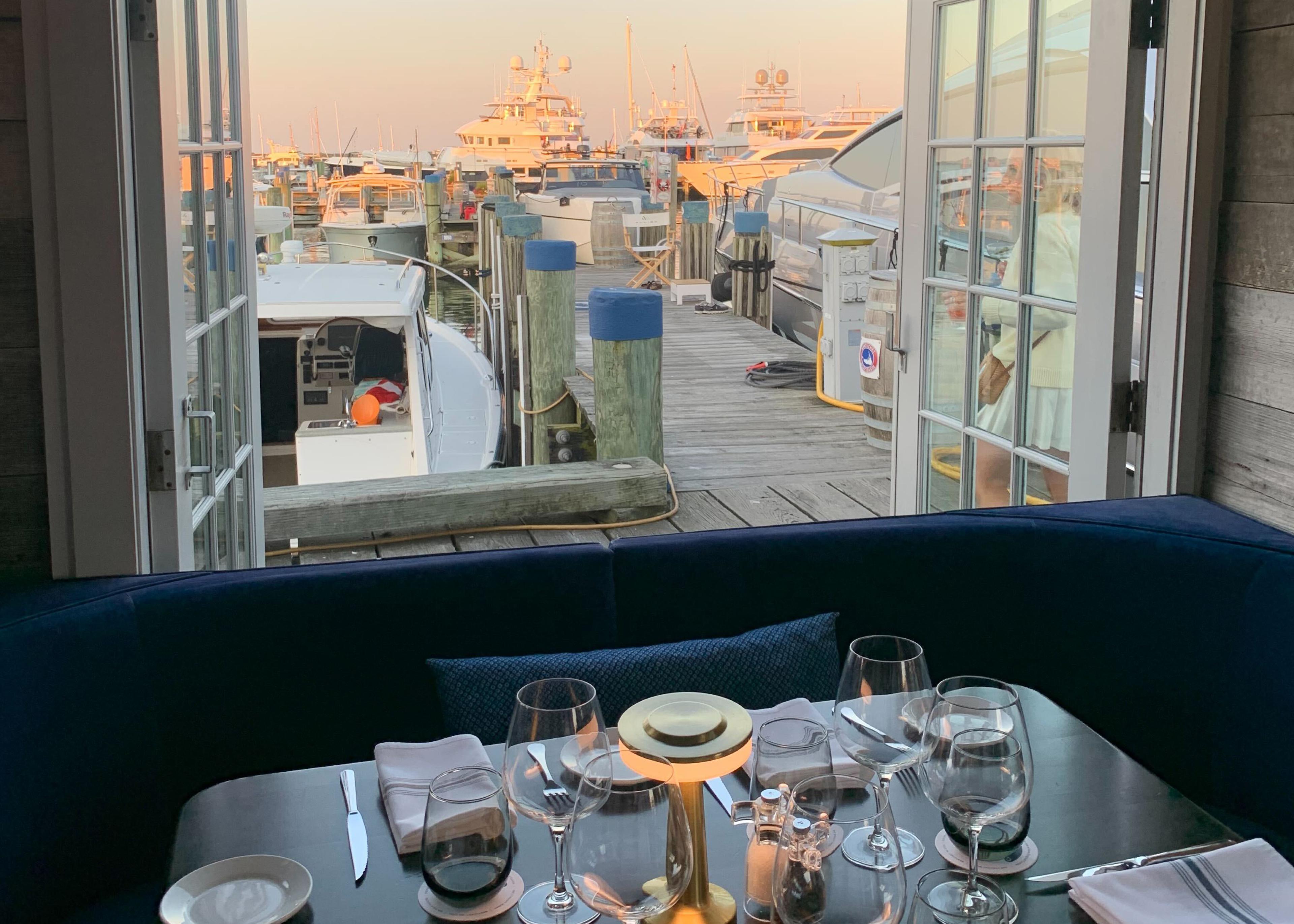 A table set for dinner at Cru with navy velvet banquette seating and open French doors framing the yacht-filled marina at dusk.