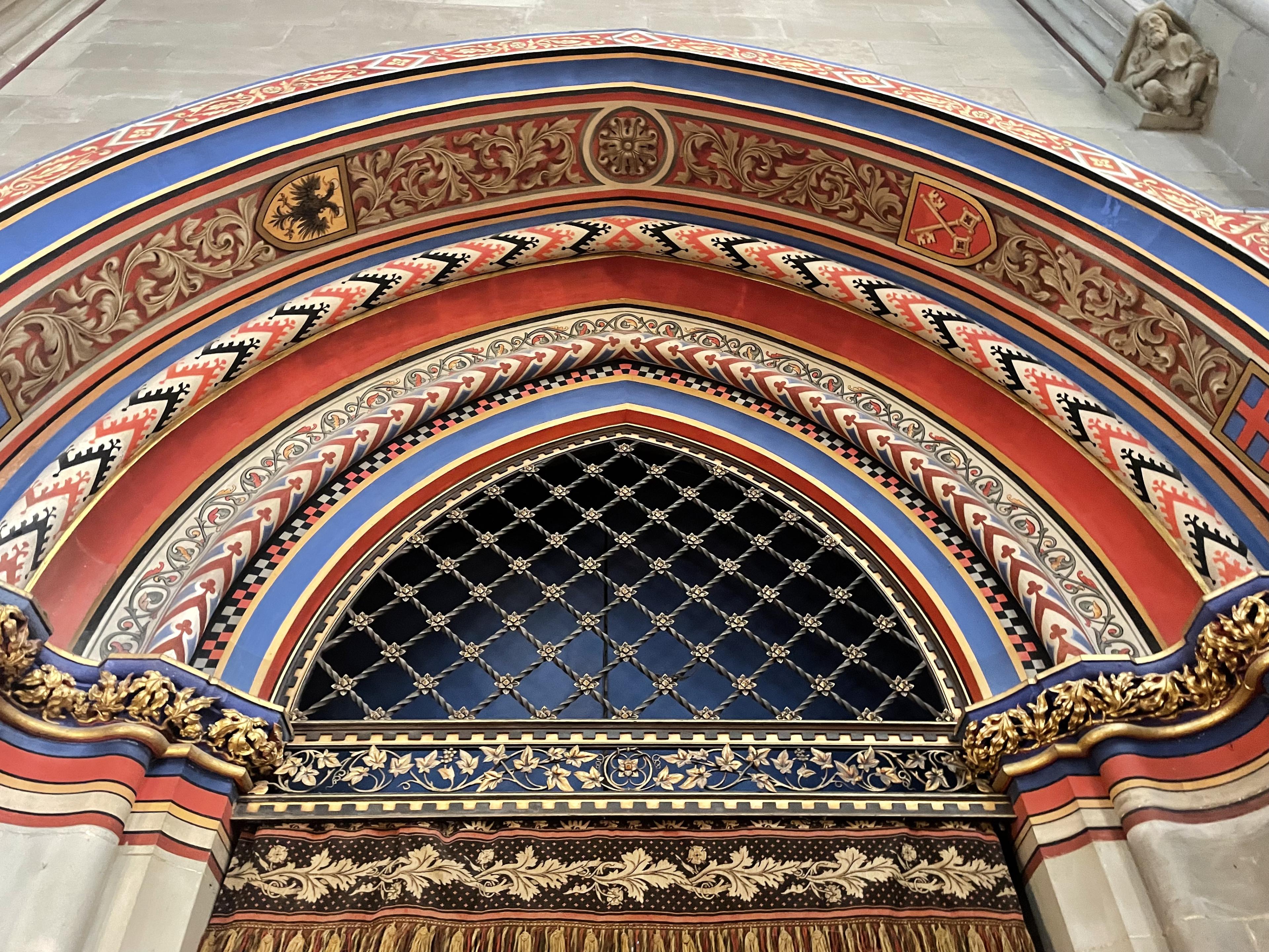 Ornate church arch with red, blue and gold patterns, heraldic shields, gilded trim, and a black lattice screen.