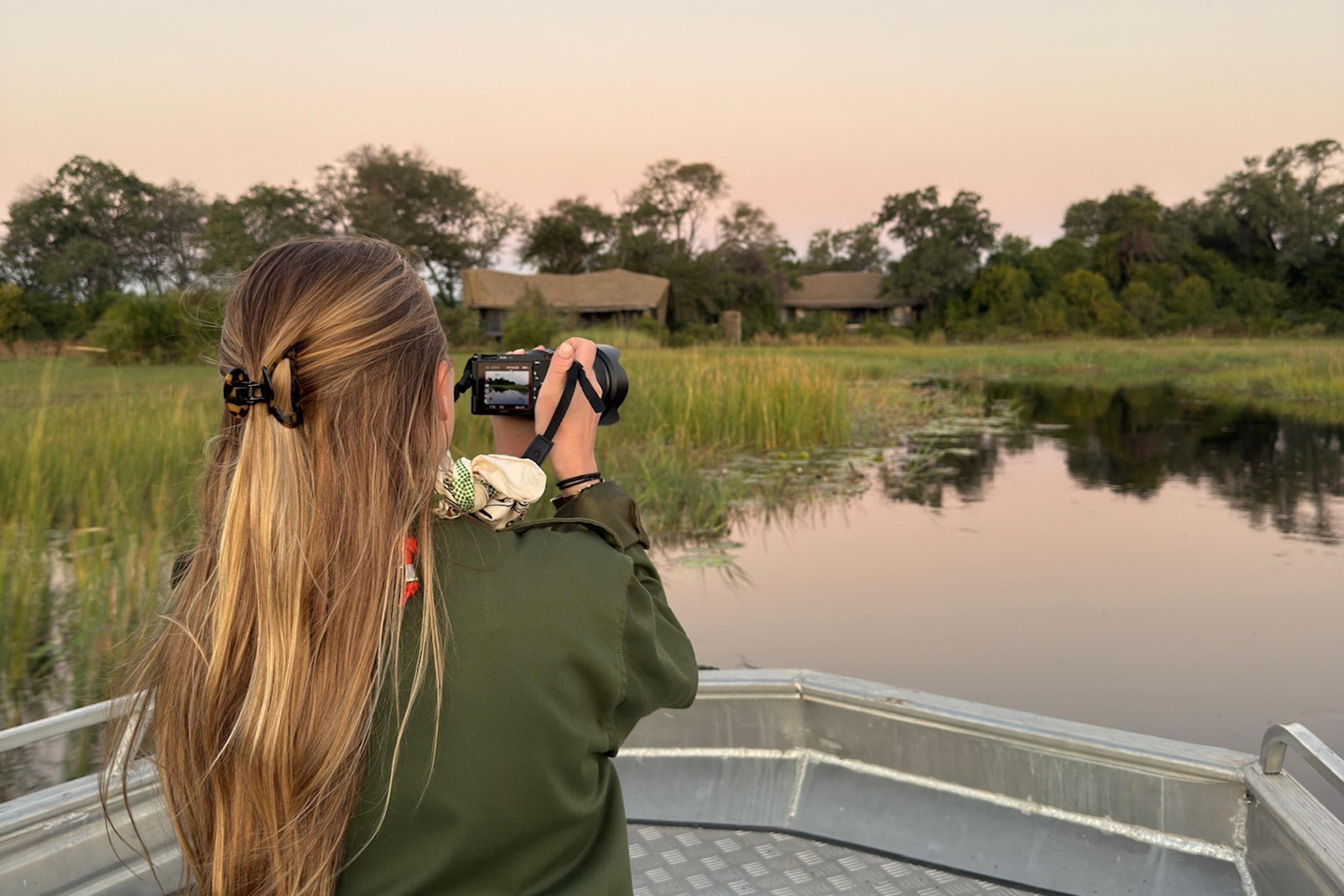 Person with long hair photographs a marshy river from a metal boat, with trees and safari lodges at sunset.