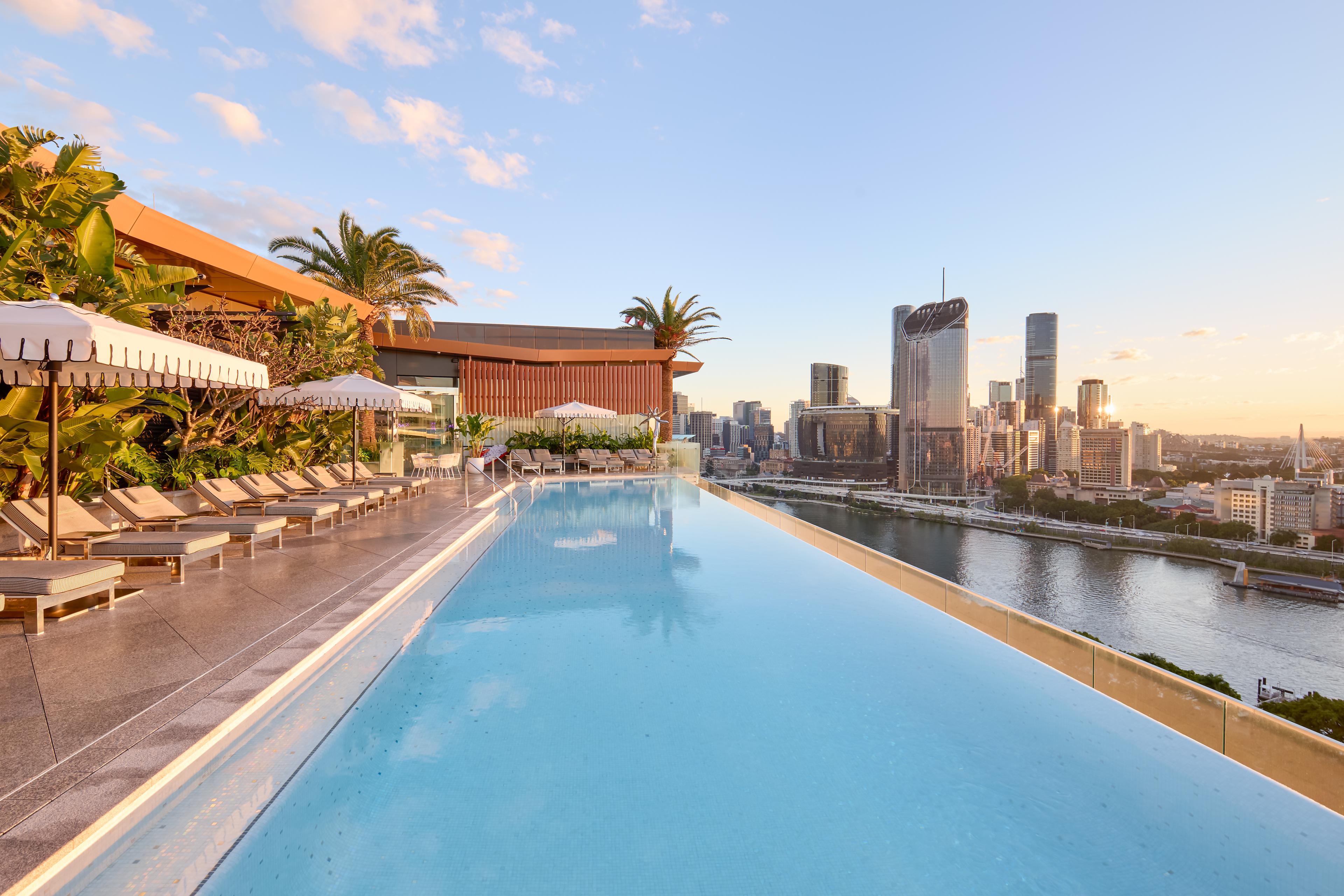 Rooftop infinity pool with lounge chairs and palms overlooking a city skyline at sunset, Emporium Hotel South Bank