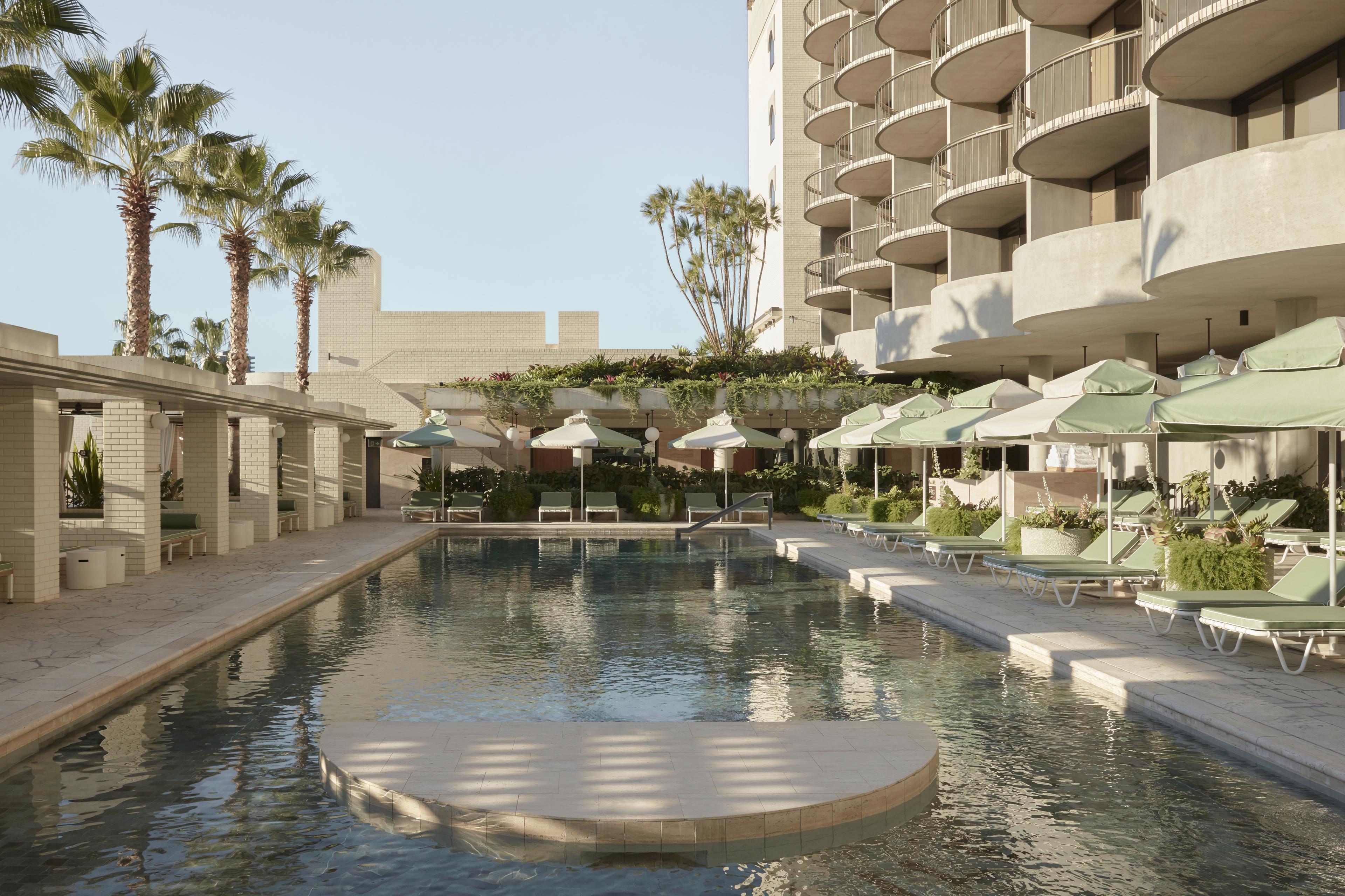 Sunlit hotel pool with palm trees, green loungers and umbrellas, surrounded by curved balconies and lush planters, The Calile