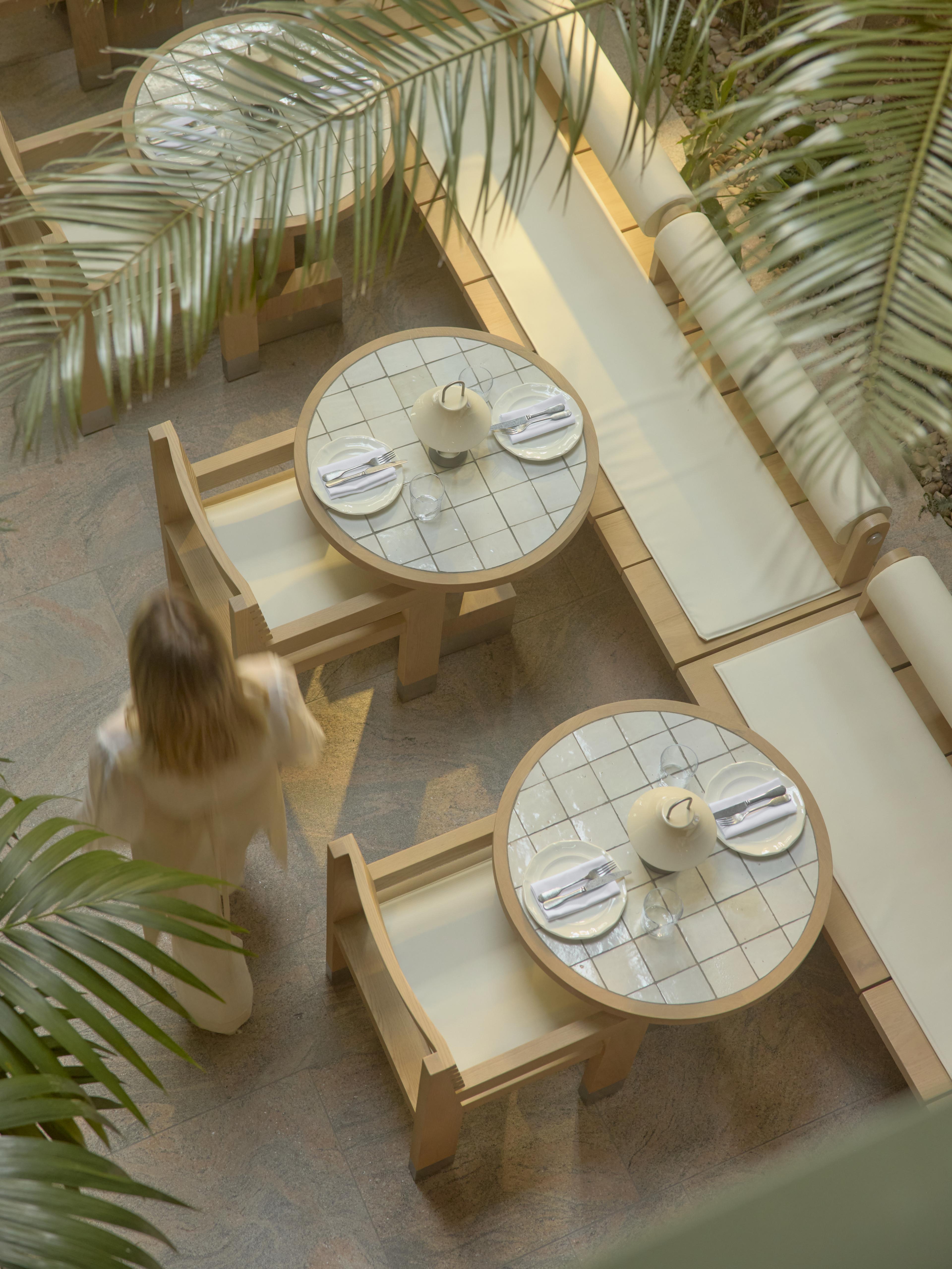 Overhead view of a leafy cafe patio with round tiled tables set for two, wooden benches, and palm fronds in the foreground, Golden Avenue