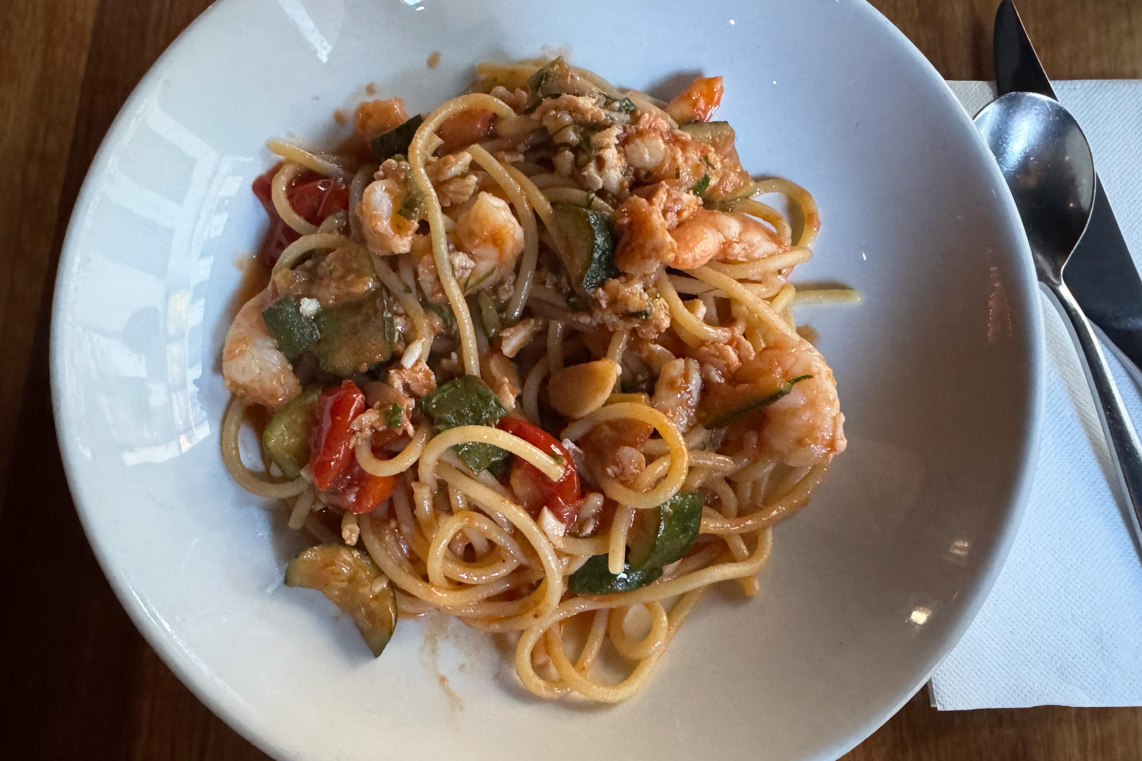 White bowl of spaghetti with shrimp, zucchini, and tomato sauce, with a spoon on a napkin beside it at Julius Pizzeria