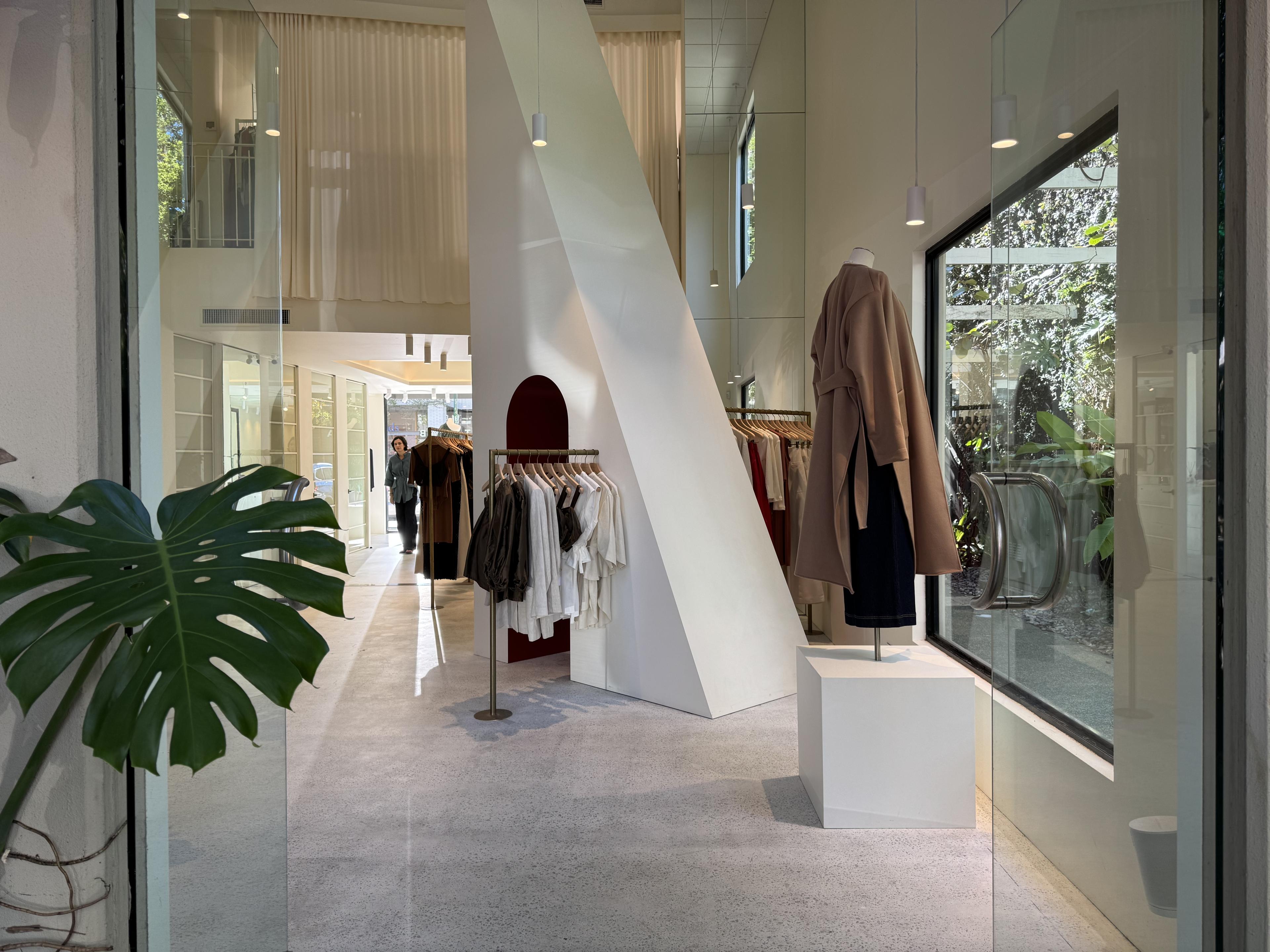 Bright boutique interior on James Street with clothing racks, a white sculptural staircase, pendant lights, and a large plant by the glass entrance.