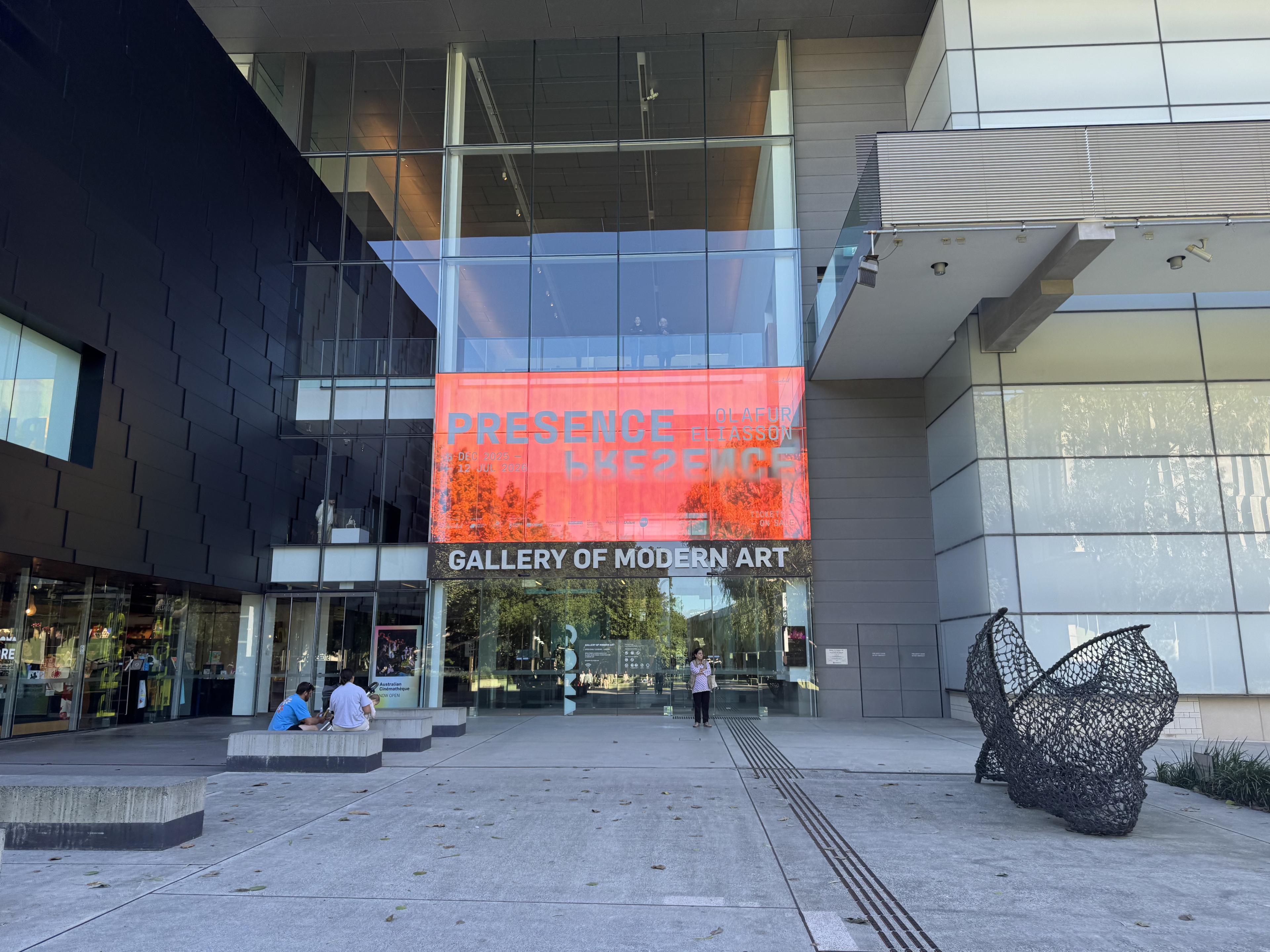 Glass-fronted Gallery of Modern Art entrance with a red exhibition banner, black wall, wire sculpture, and people seated outside.