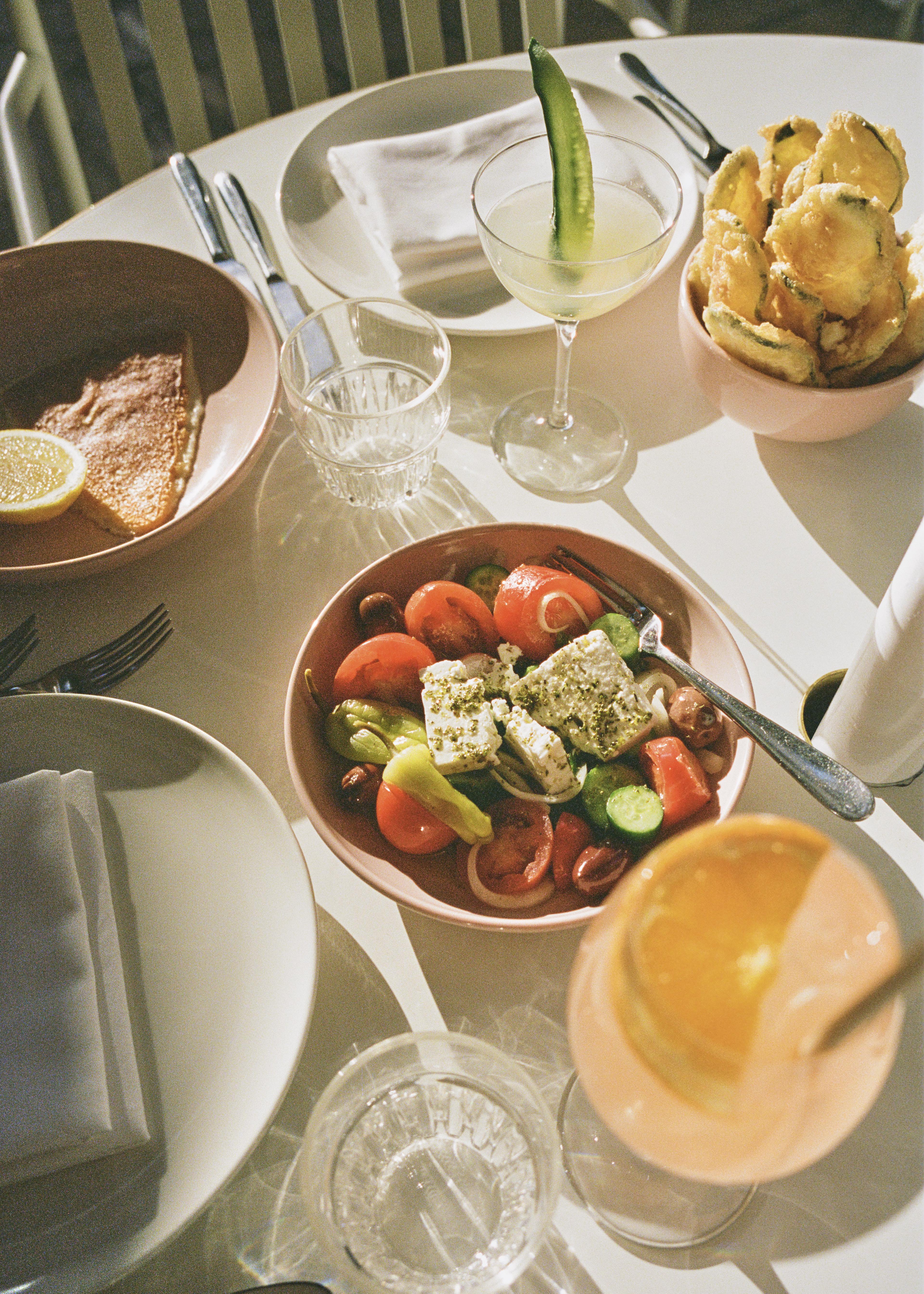 Sunlit table with salad, chips, fish, drinks, plates, cutlery, and napkins at Hellenika