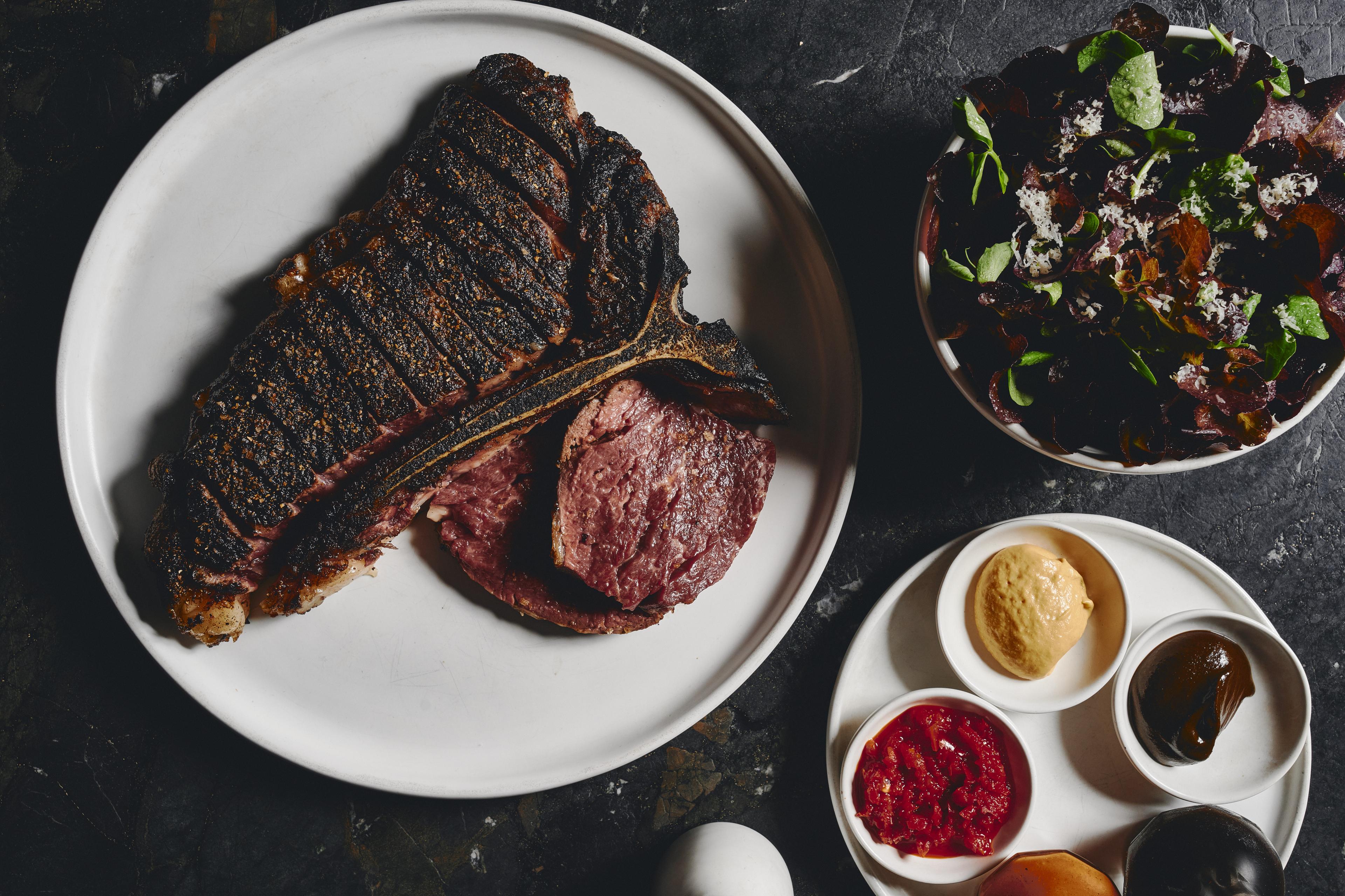 Sliced grilled steak on a white plate with greens, grated cheese, and small bowls of sauces at Agnes.