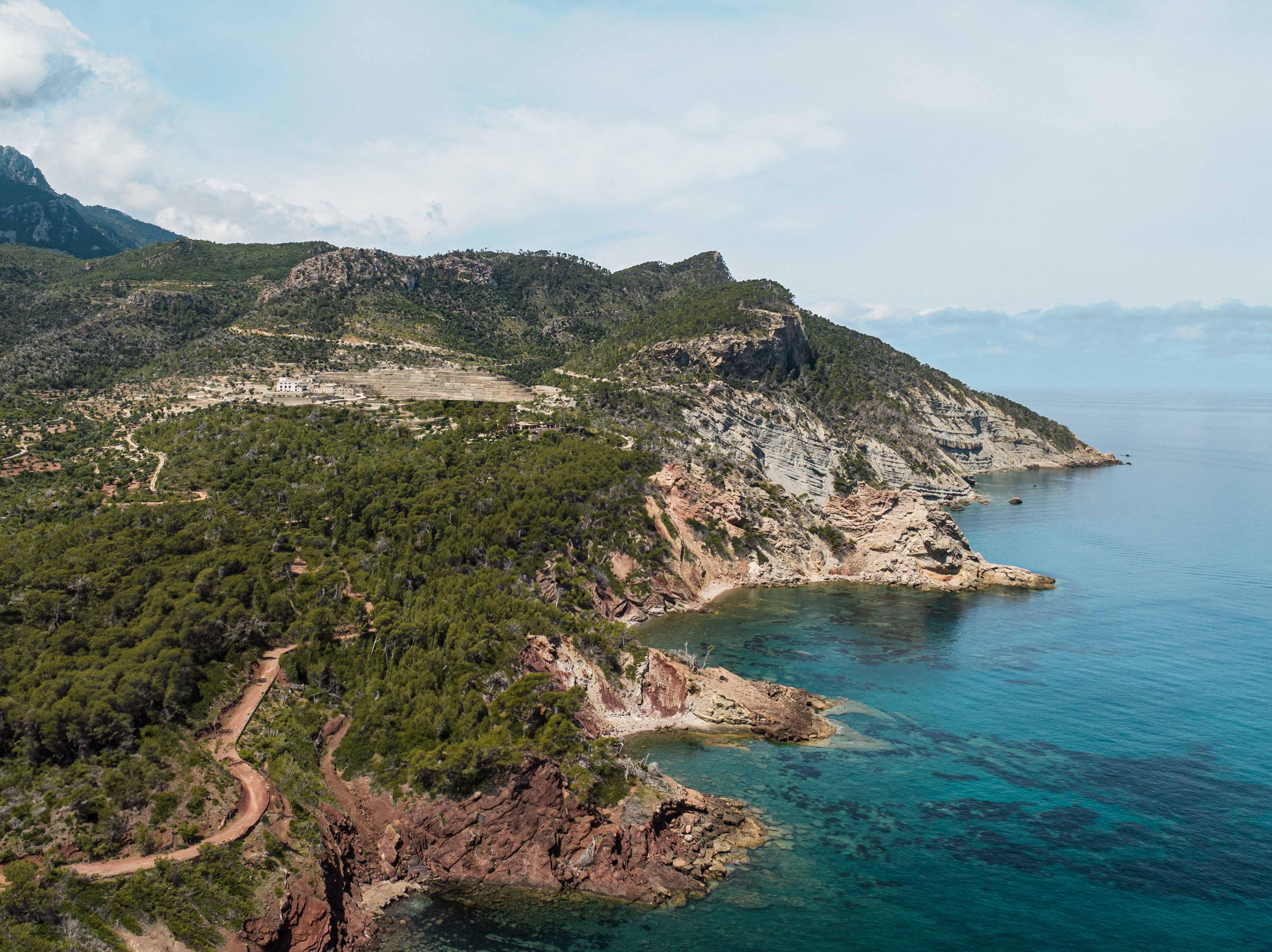 Rocky green coastline with winding dirt road, terraced hills, cliffs, and clear turquoise sea under a pale sky in Mallorca