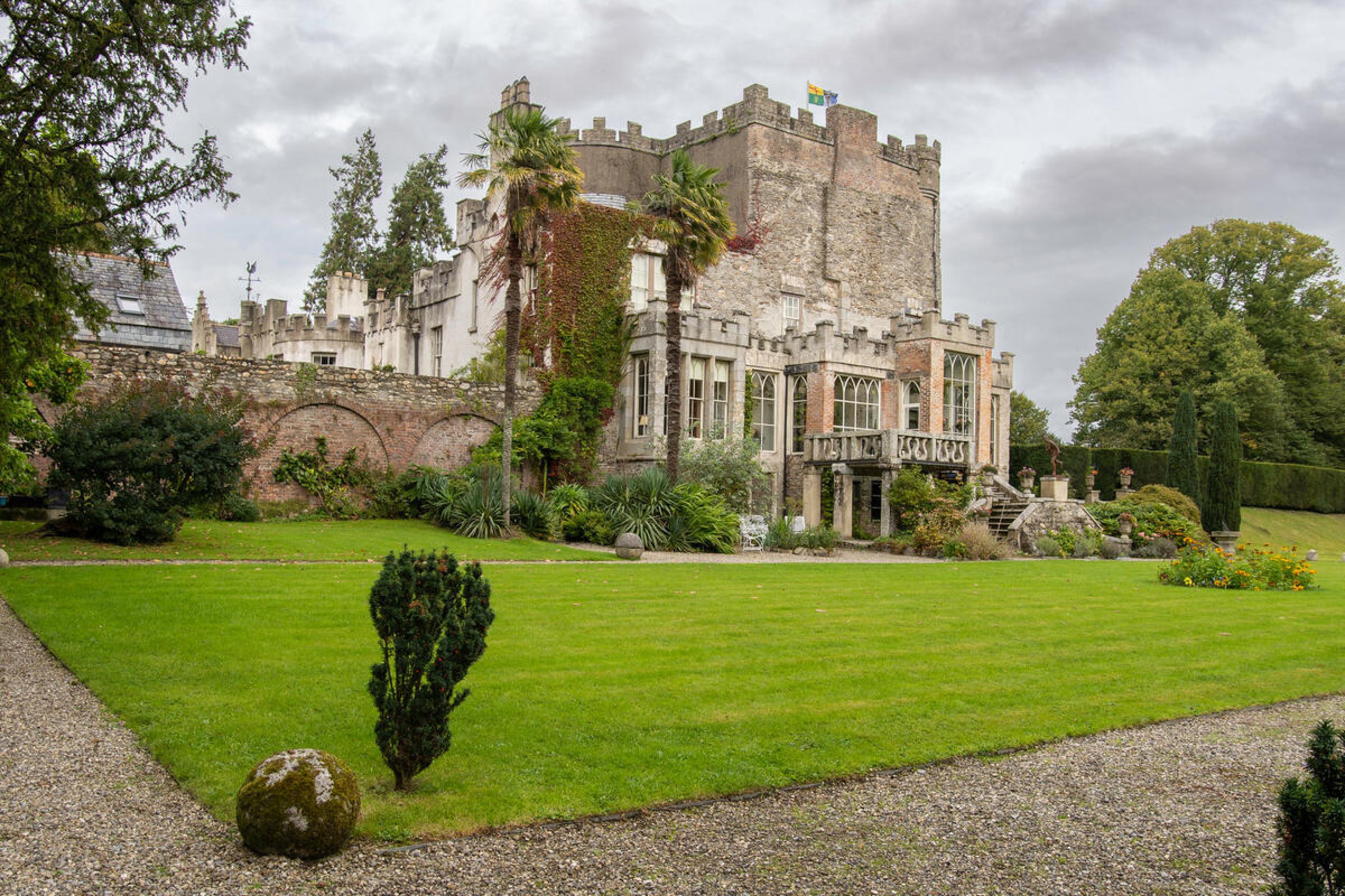 Stone castle with ivy and battlements overlooking a manicured lawn and garden under a cloudy sky.