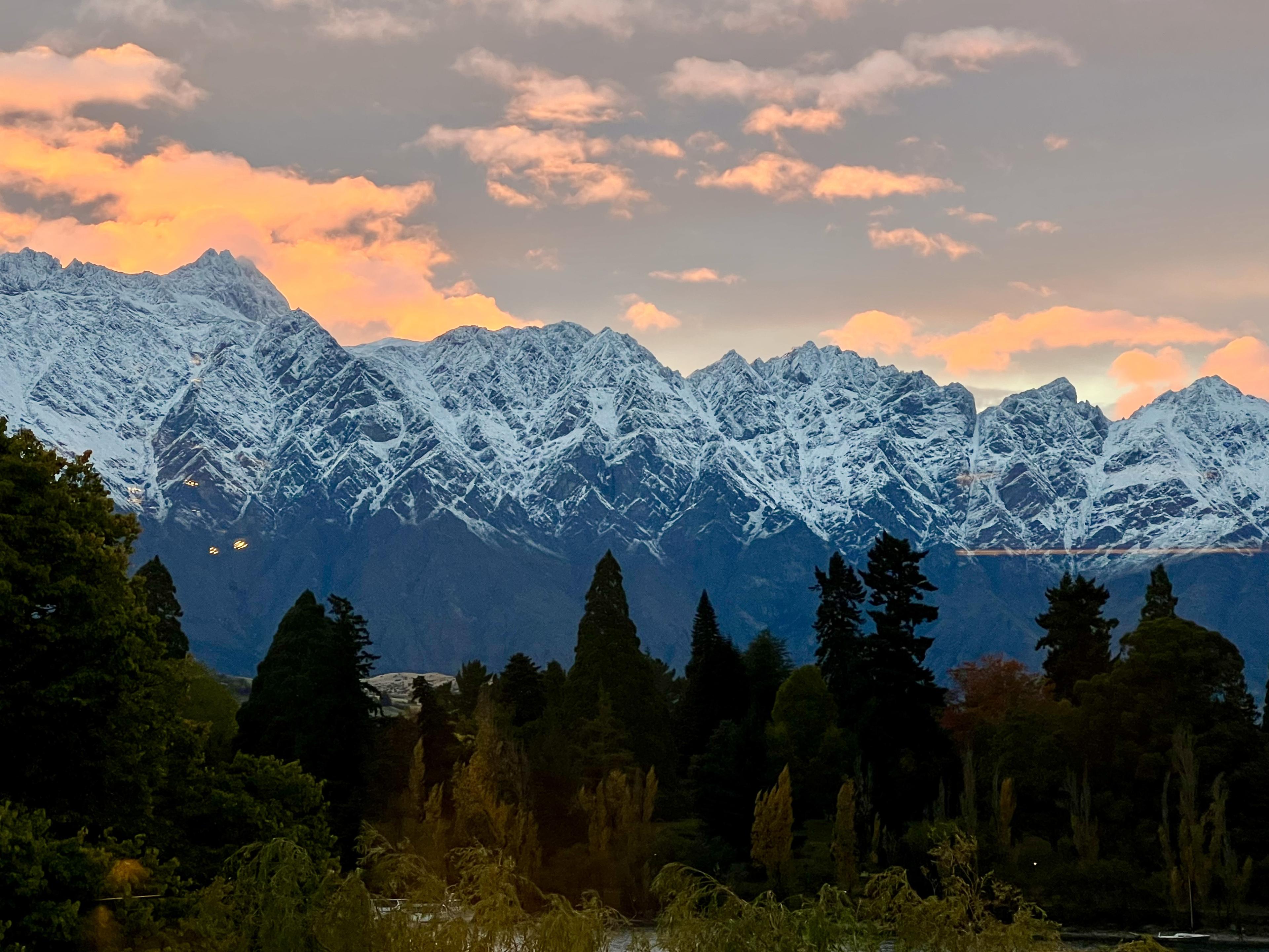 Snowy mountain range beneath peach sunset clouds, with dark trees in the foreground.