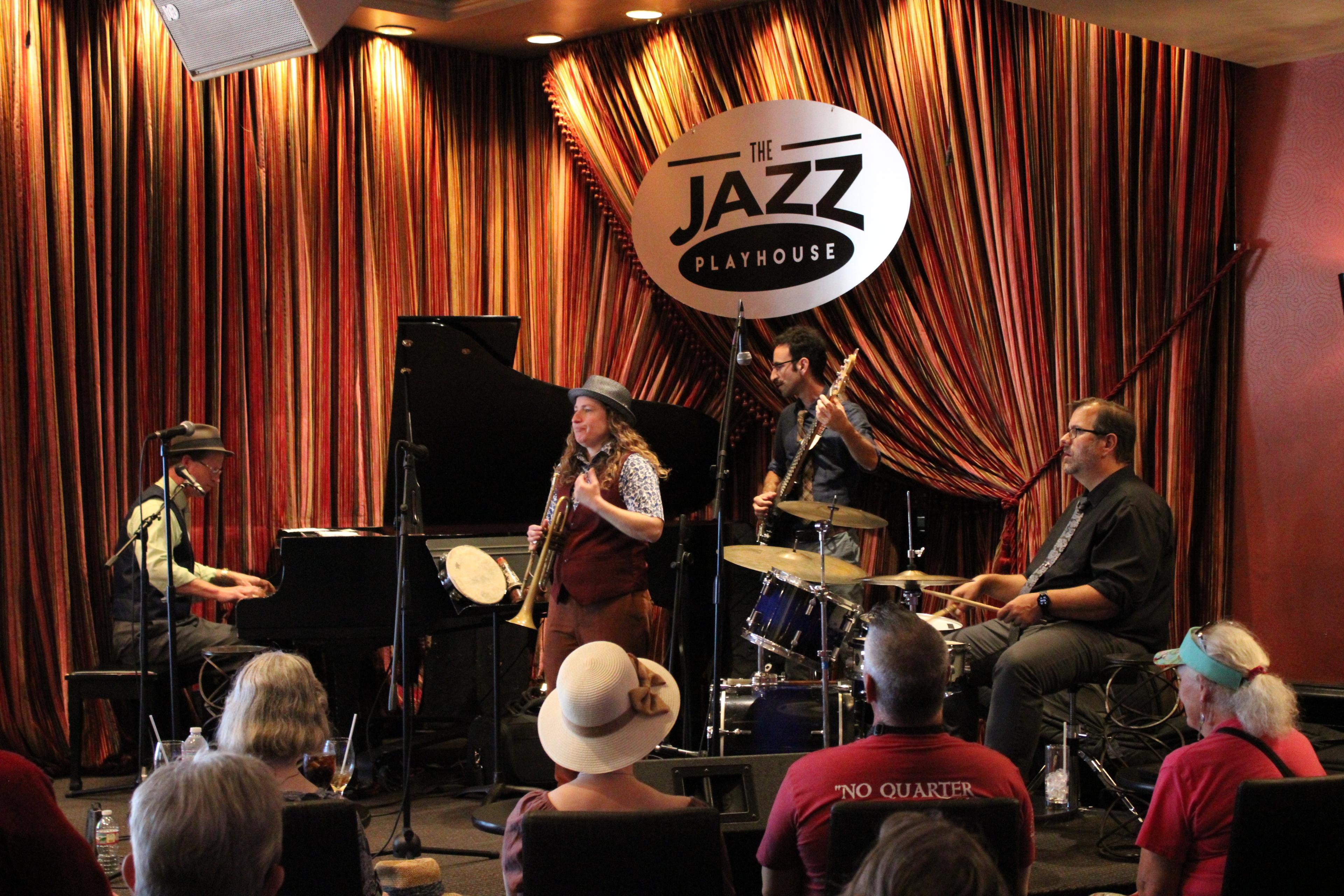 Jazz band performs at The Jazz Playhouse before striped curtains, with piano, brass, drums, and audience in foreground.