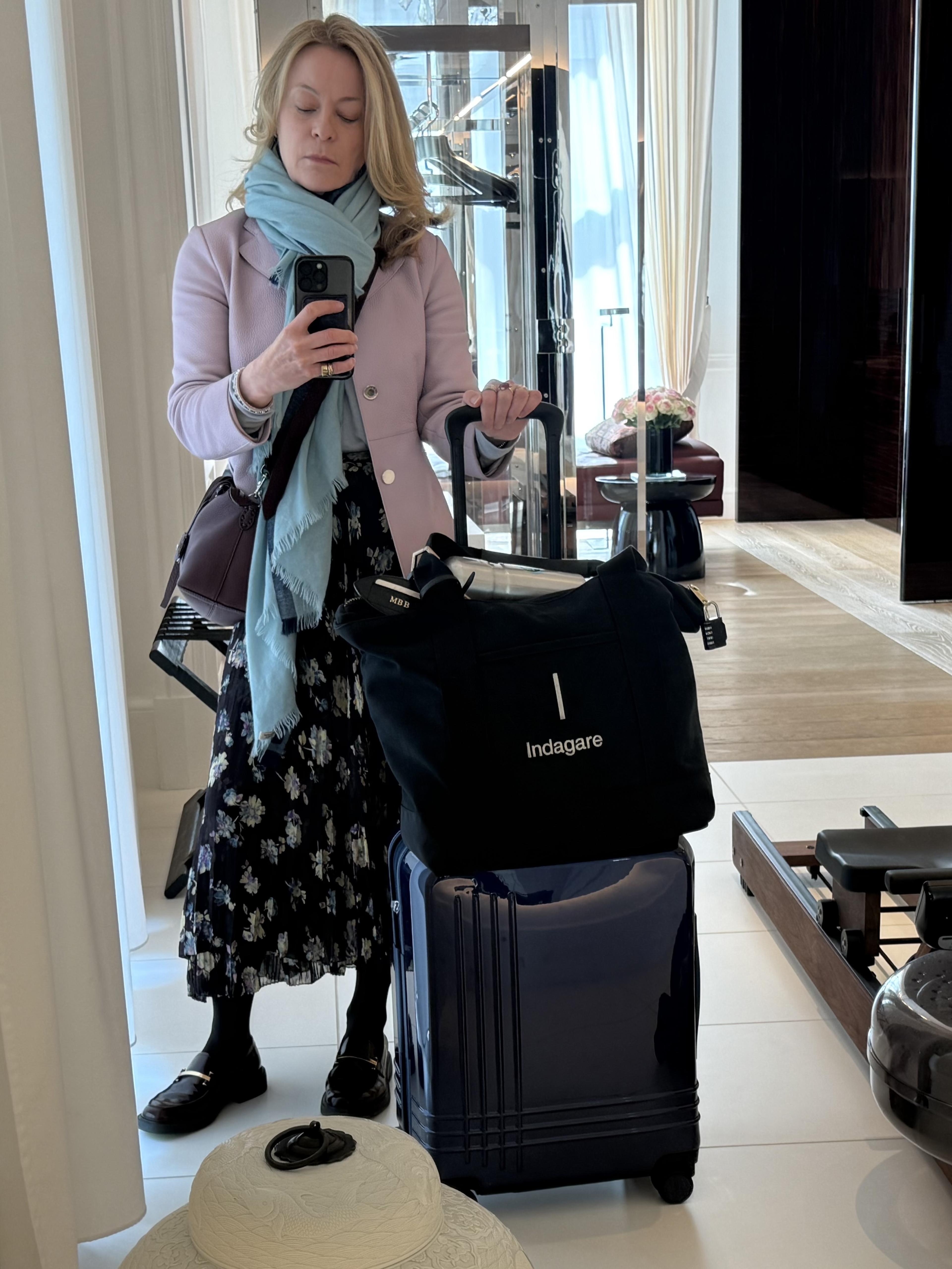 Woman in pastel coat and blue scarf takes a mirror selfie beside stacked luggage in a bright hotel room.
