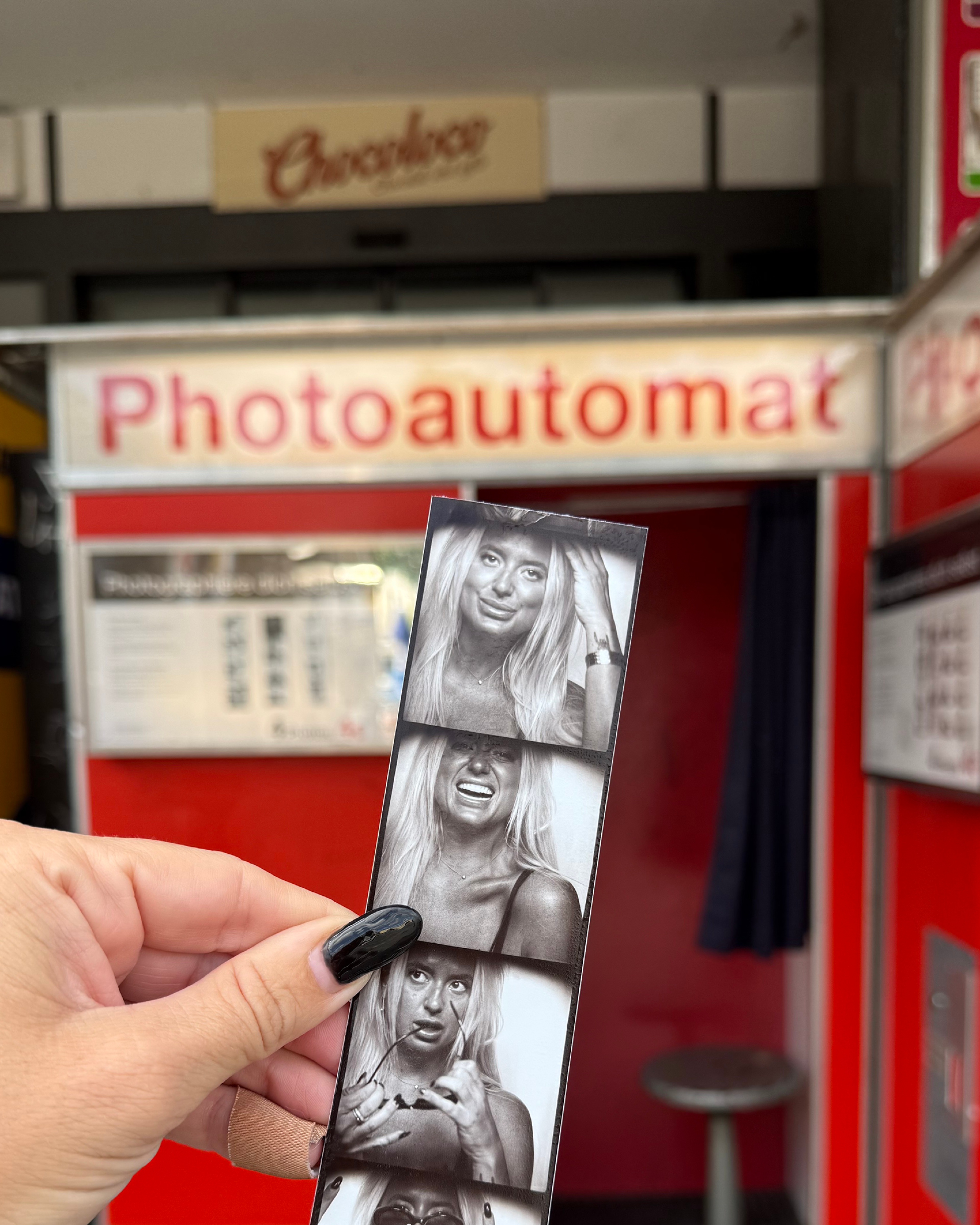 Hand holding a black-and-white photobooth strip of a blonde woman in front of a red Photoautomat booth.