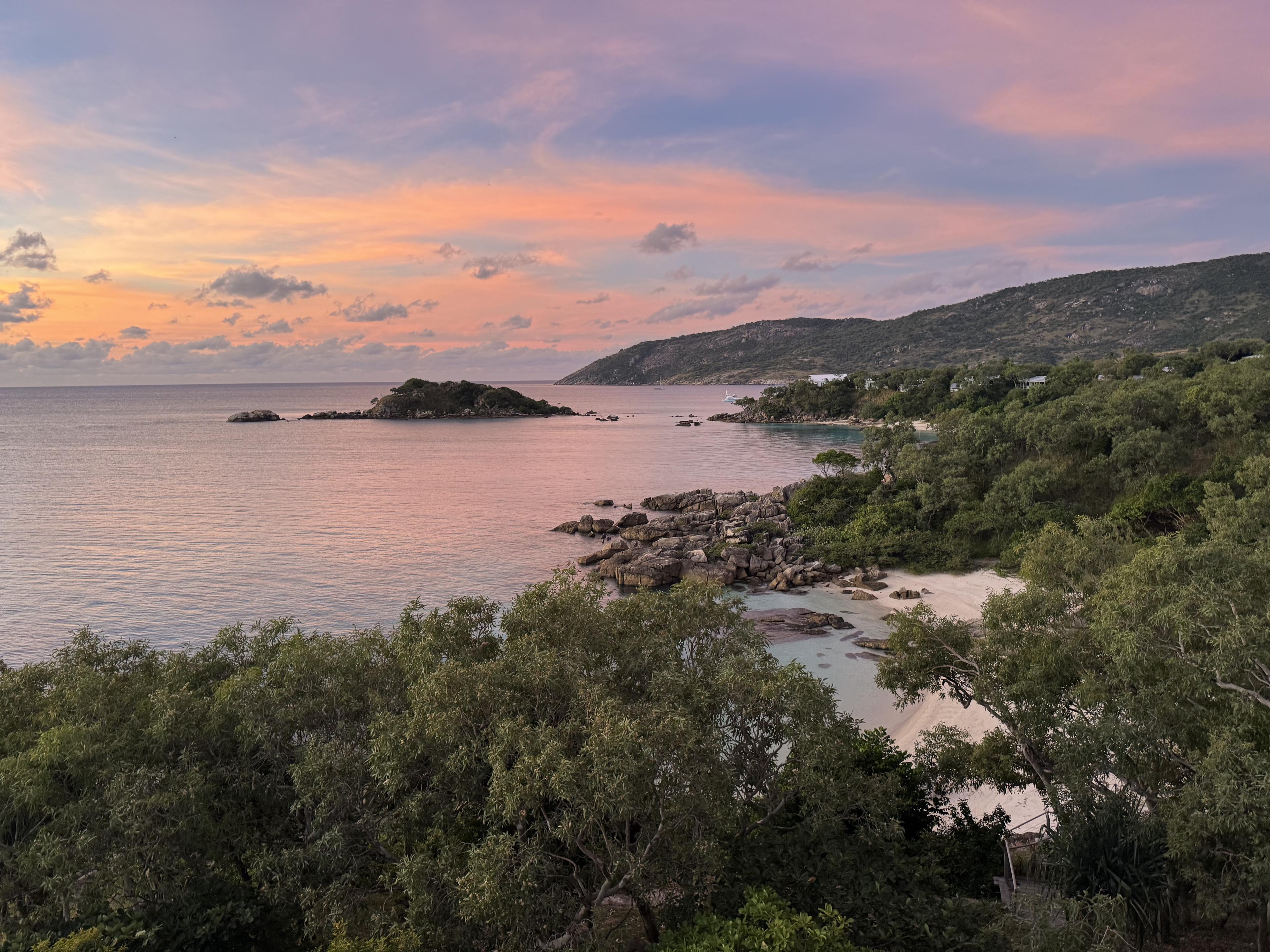 Pink-orange sunset over a calm coastal bay, with rocky hills, small boats, sandy coves, and trees in the foreground.