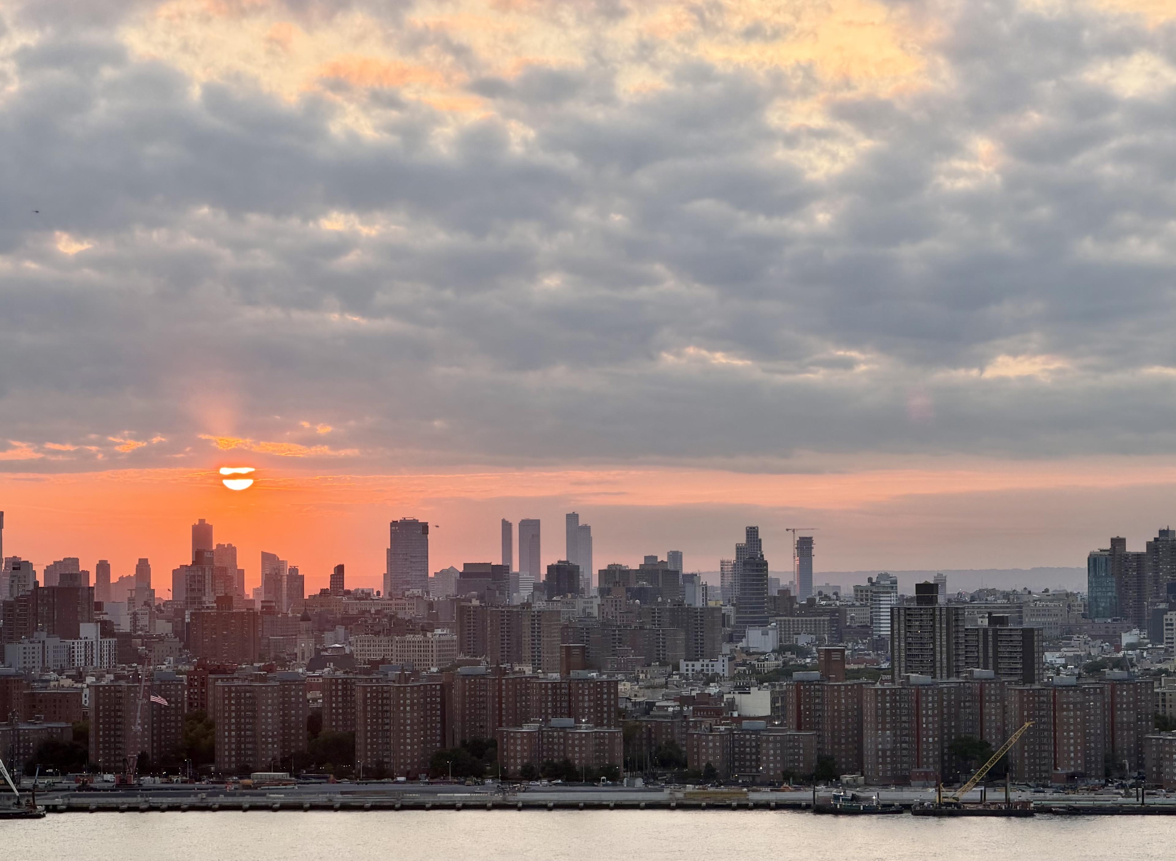 Orange sunset glows beneath layered clouds over a city skyline, with high-rises and a river in the foreground.