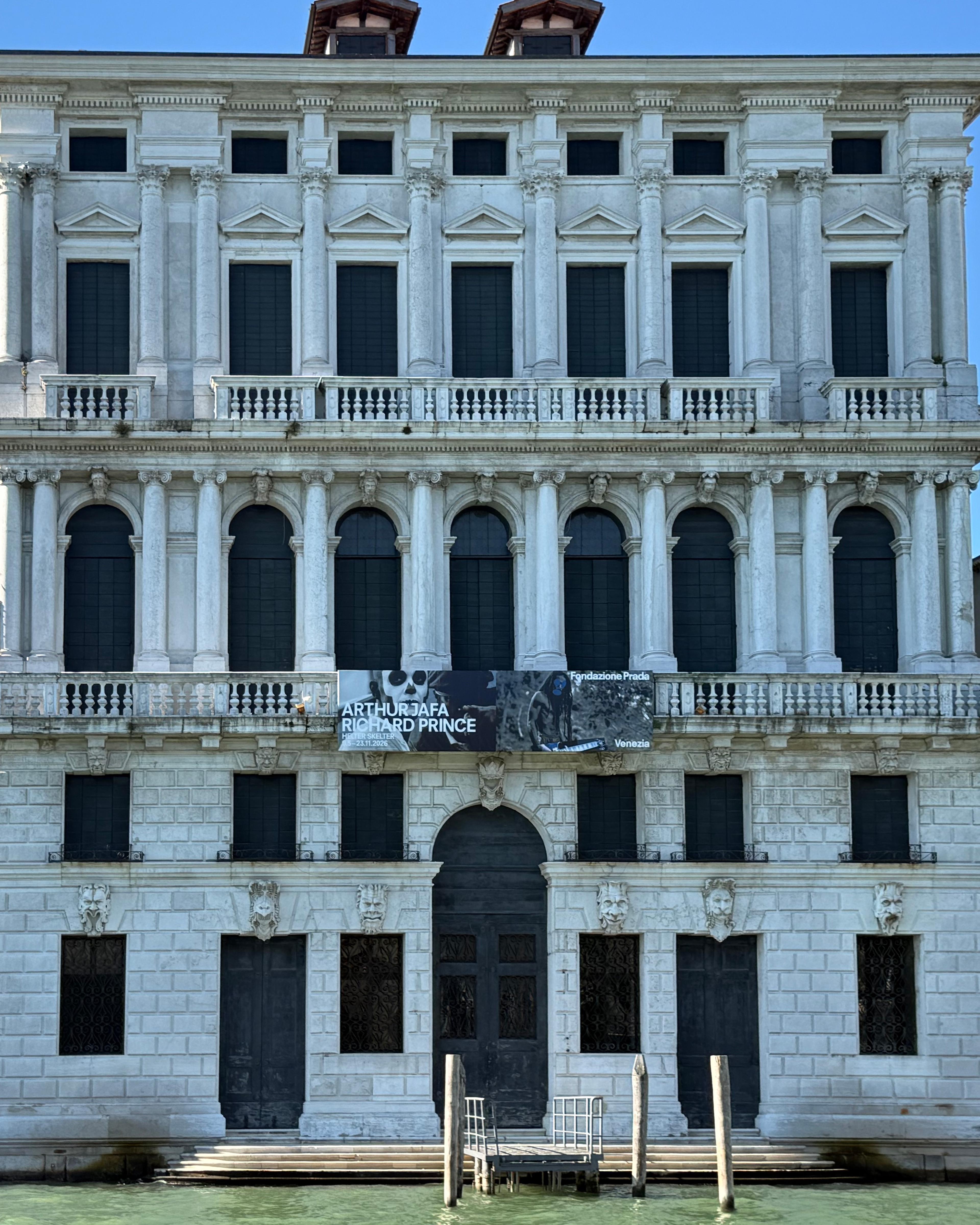 Ornate Venetian palazzo facade with arched windows, columns, canal steps, and a Fondazione Prada exhibition banner.