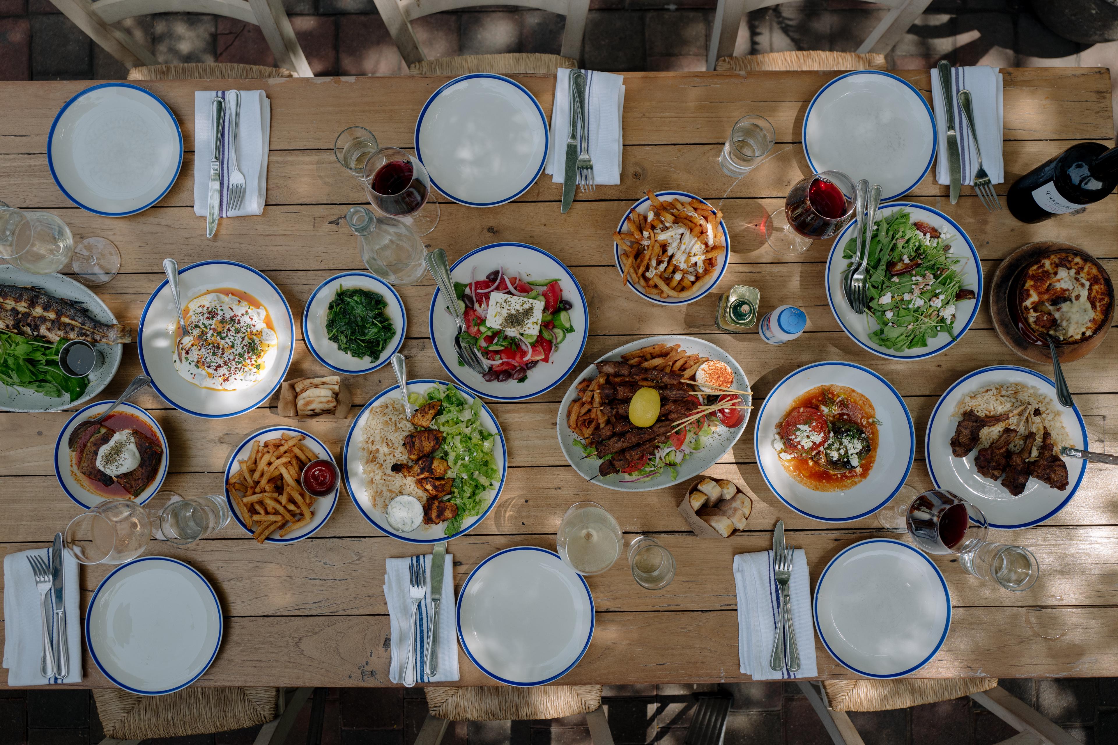 Overhead view of a wooden table set with blue-rimmed plates, cutlery, wine, and assorted Mediterranean dishes.