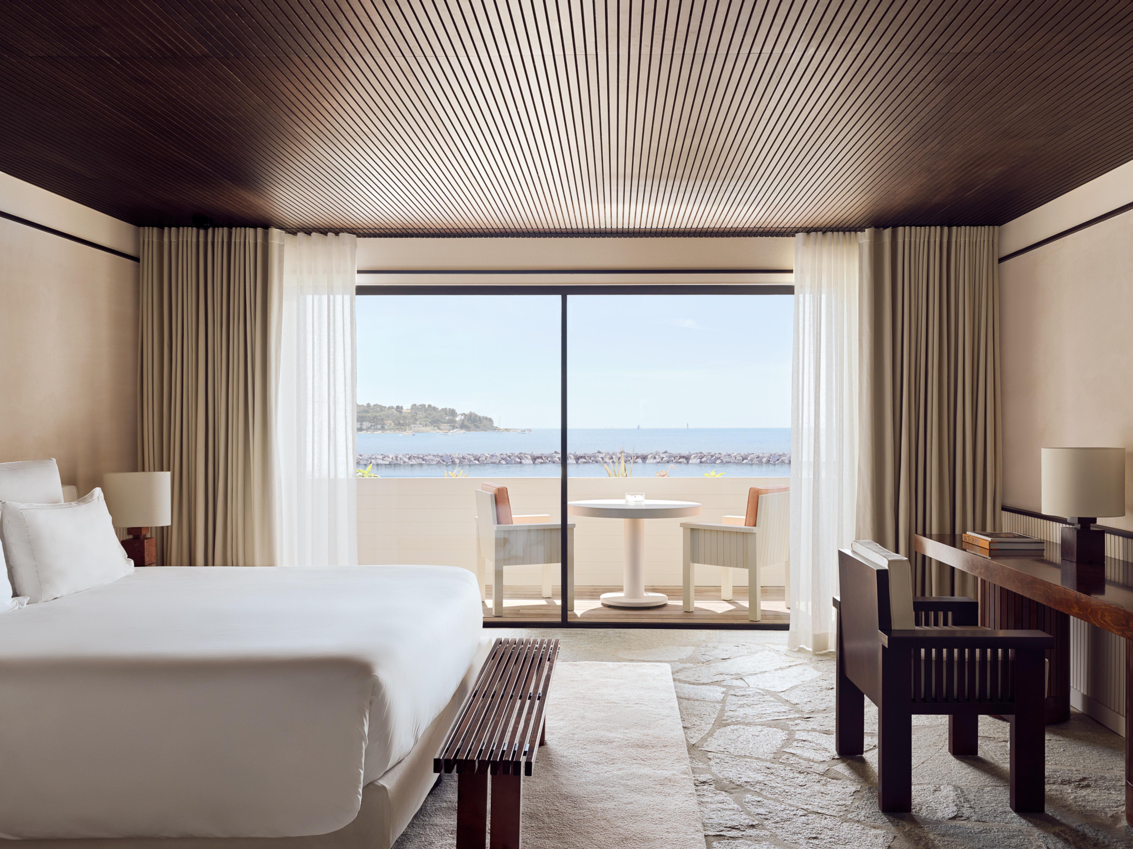 Bed with white linens on a stone floor and a small wooden desk opposite the bed, all overlooking a small patio and the sea 